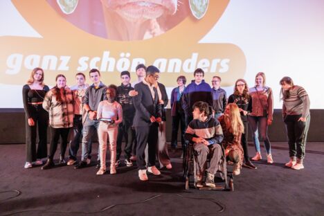 15 Schüler*innen und Lehrkräfte stehen auf der Bühne des Astor Kinos und blicken in die Kamera. Ein Junge sitzt im Rollstuhl.