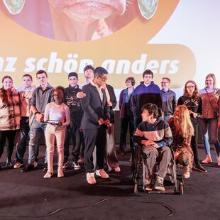 15 Schüler*innen und Lehrkräfte stehen auf der Bühne des Astor Kinos und blicken in die Kamera. Ein Junge sitzt im Rollstuhl.
