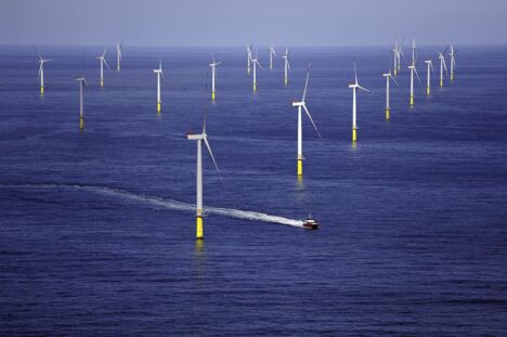 Windpark in der Nordsee. Dutzende Windräder drehen sich. Ein Schiff fährt mit hoher Bugwelle an den Windrädern vorbei.